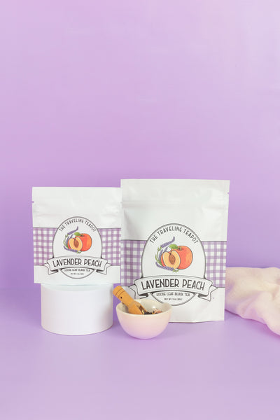 One small and one large retail package of lavender peach black tea next to a cup of loose tea leaves on a purple background
